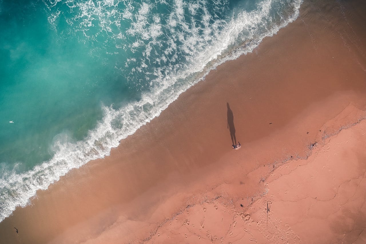 A bird's eye view of a beach with water and waves viewed from southwest to northeast. A small figure stands on the beach, but casts a noticeable shadow